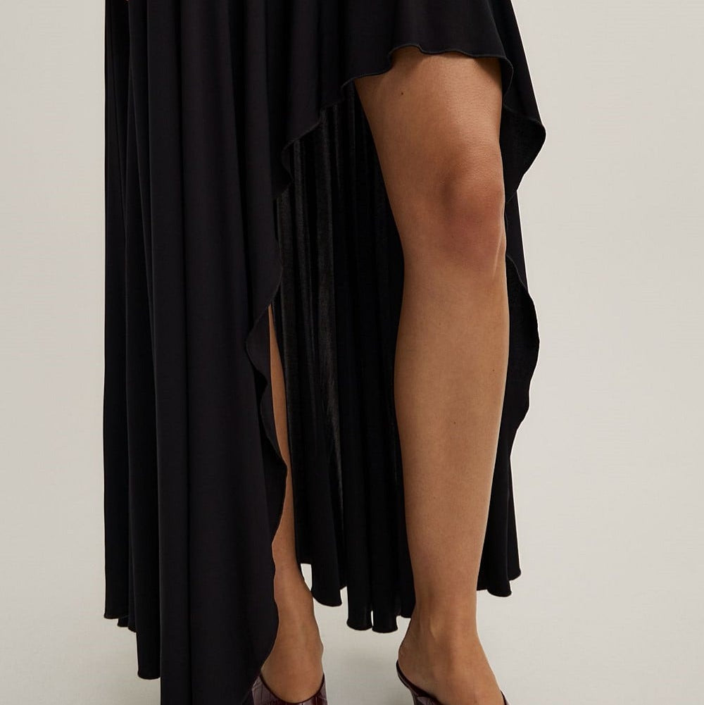 A black high-low ruffled skirt with a side slit, shown on a model wearing dark brown heeled thong sandals against a light gray studio background.