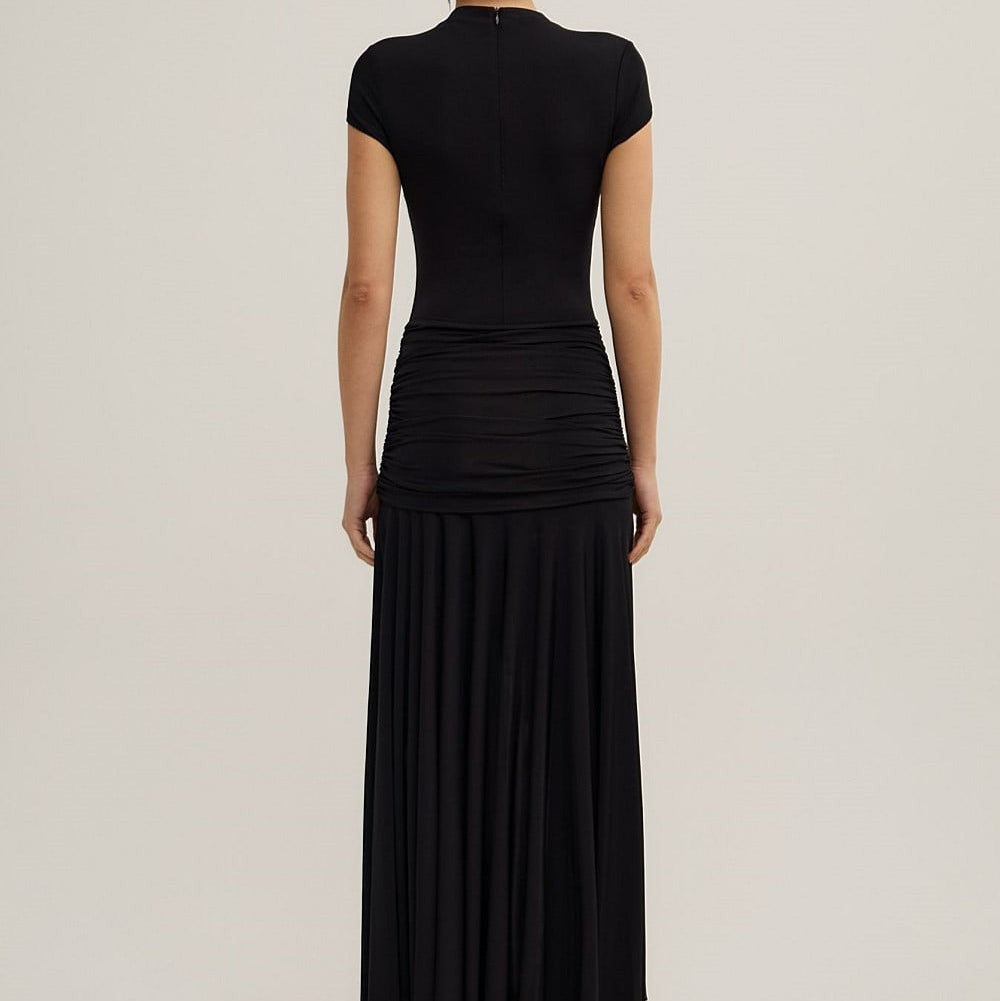 A woman viewed from the back wearing a black short-sleeve mock-neck maxi dress with a gathered waist and pleated skirt, paired with black heels, against a light gray studio background.