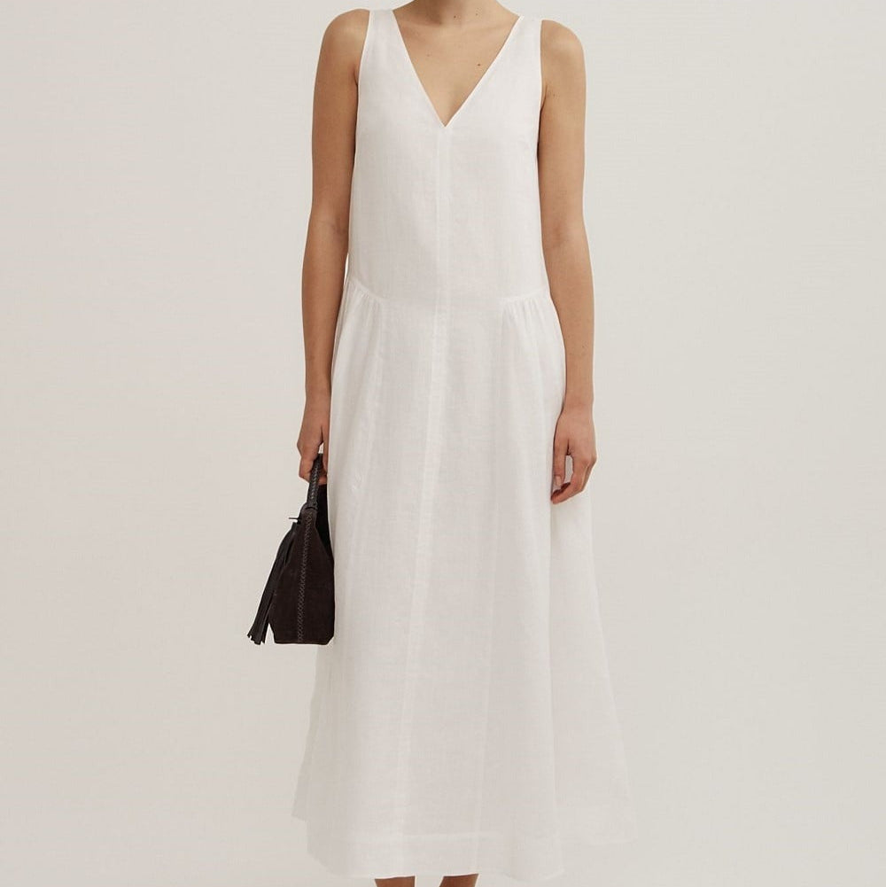 A woman modeling a sleeveless white maxi dress with a V-neckline, standing against a plain light gray studio background and holding a small black handbag.
